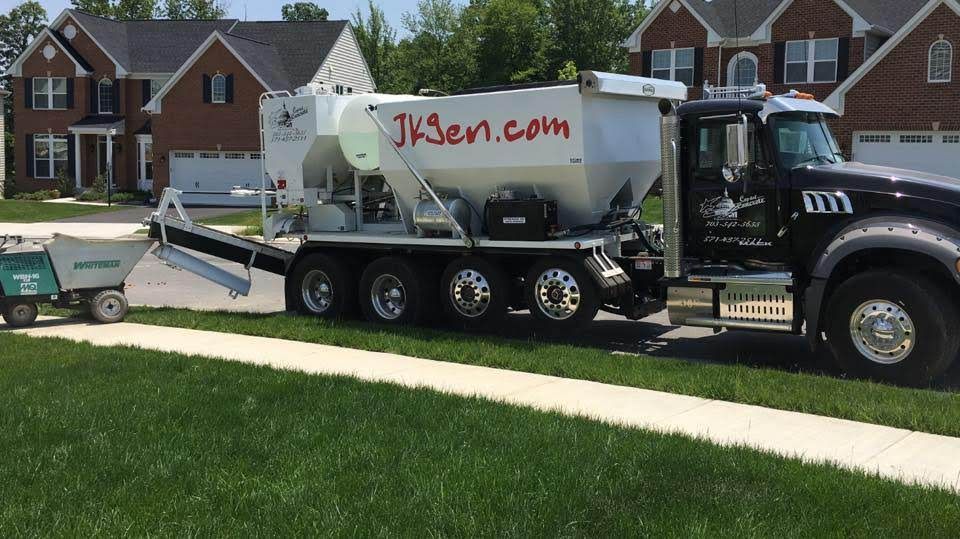 Black dump truck hauling a silver equipment trailer on a suburban street, with houses and lawn behind.
