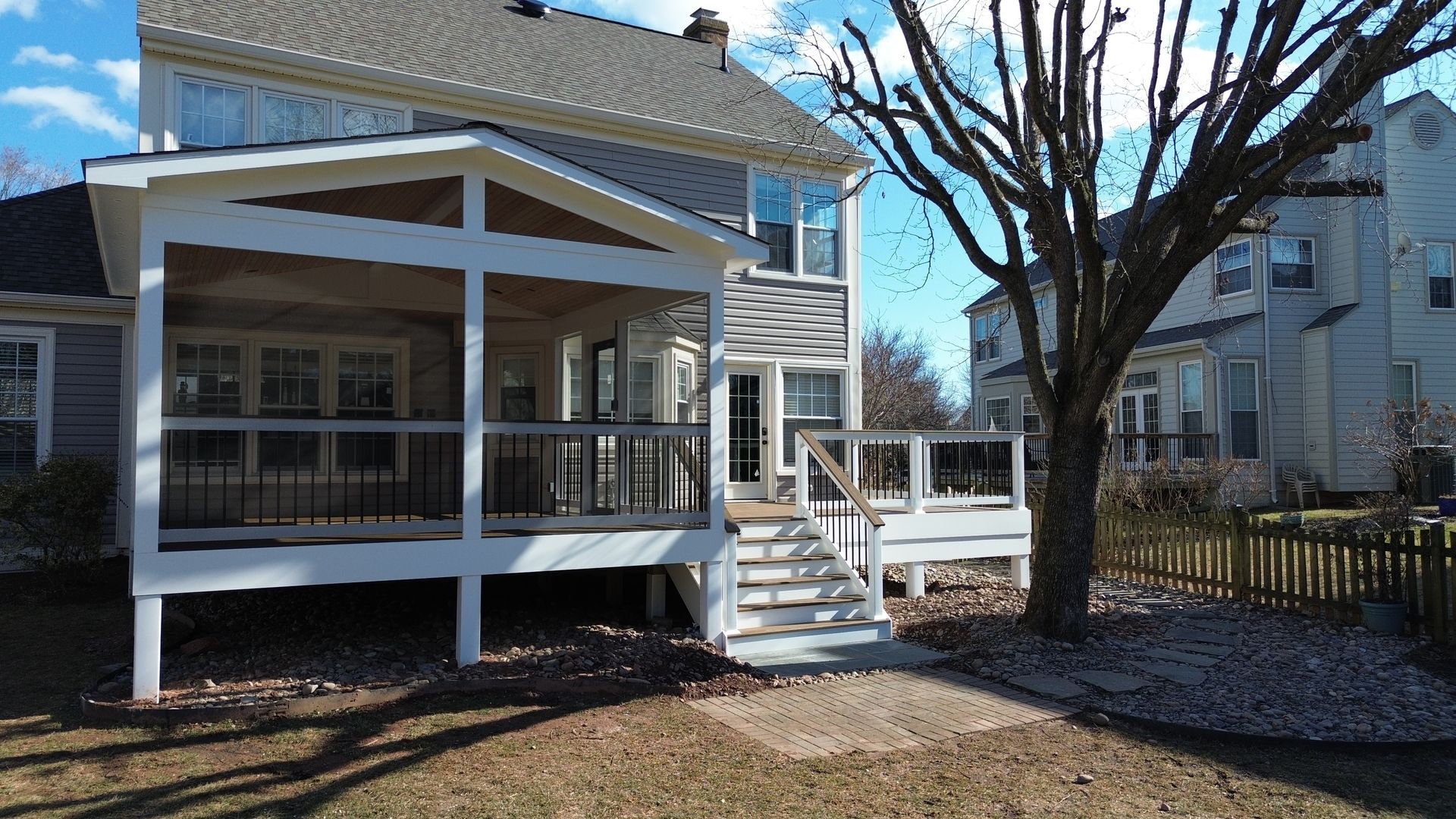 White porch attached to a two-story house with stairs, a large tree, and a fenced yard in bright daylight
