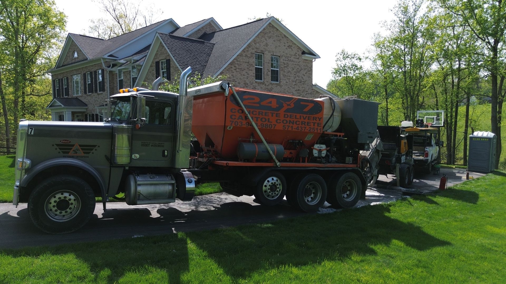 Green dump truck hauling orange equipment on a grassy driveway beside a stone house and trees