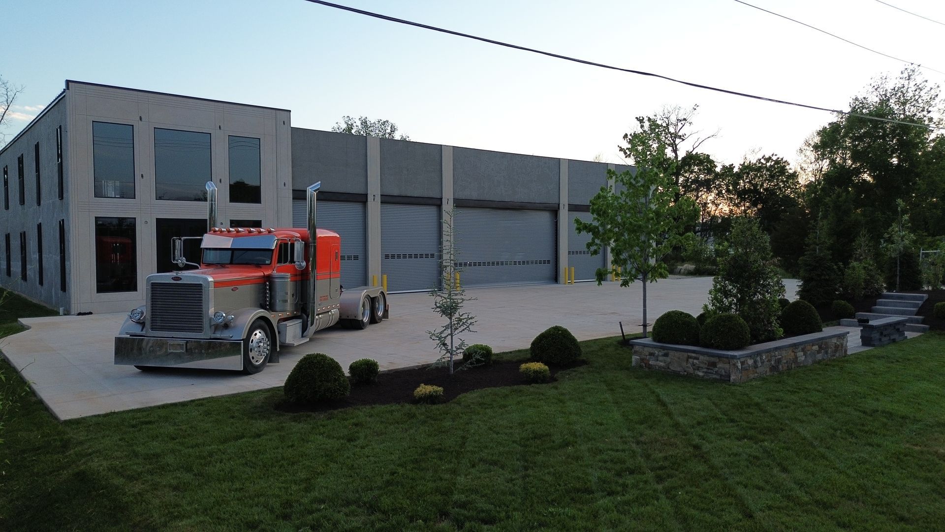 Red semi truck parked by a gray industrial building with garage doors at dusk
