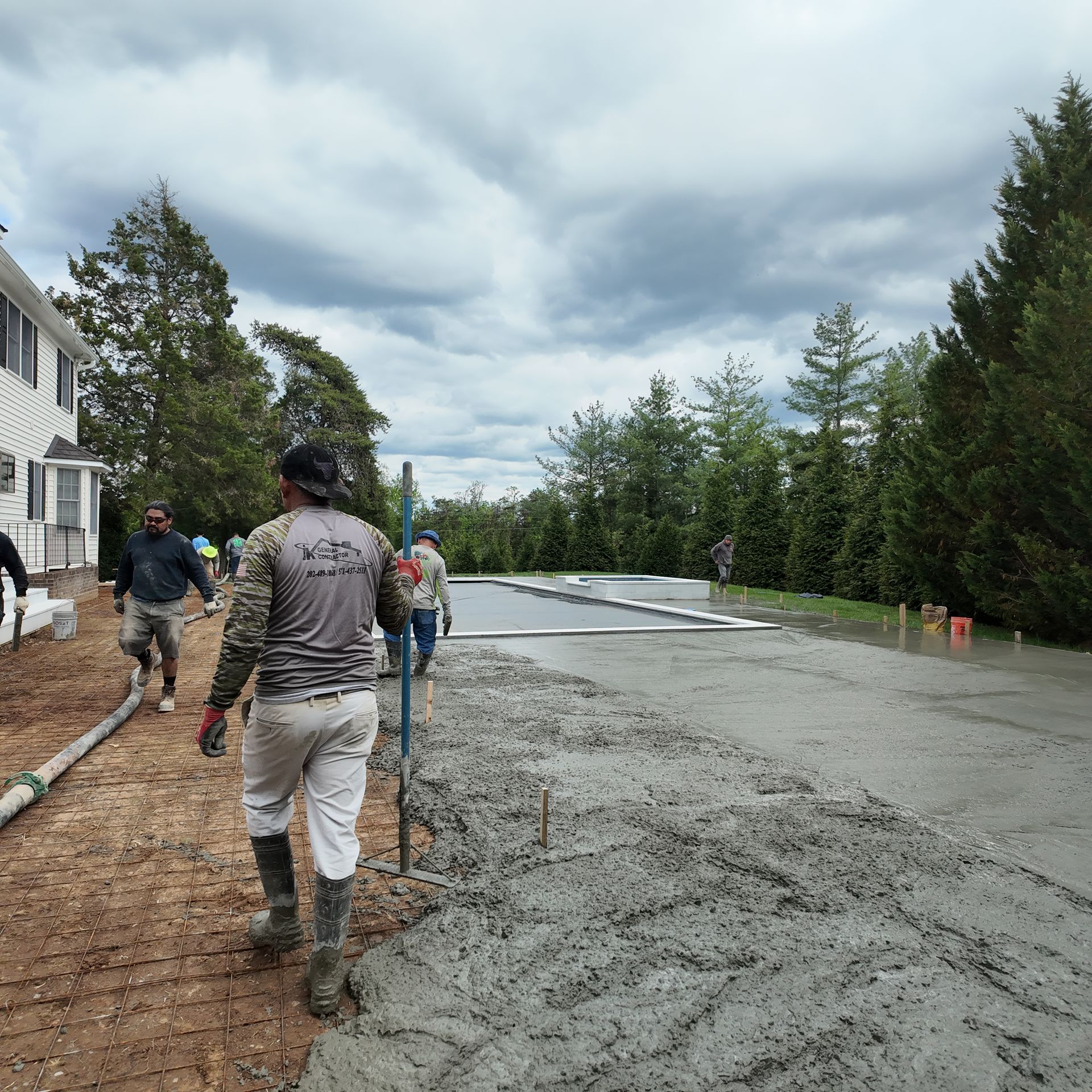 Workers spread gravel on a driveway beside a house under cloudy skies, with trees and a wet road nearby