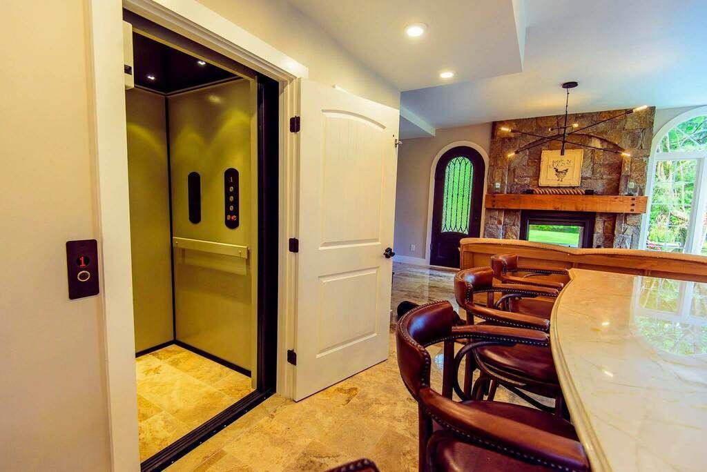 Open elevator beside a bright kitchen with white cabinets, wood bar stools, and stone fireplace backsplash.