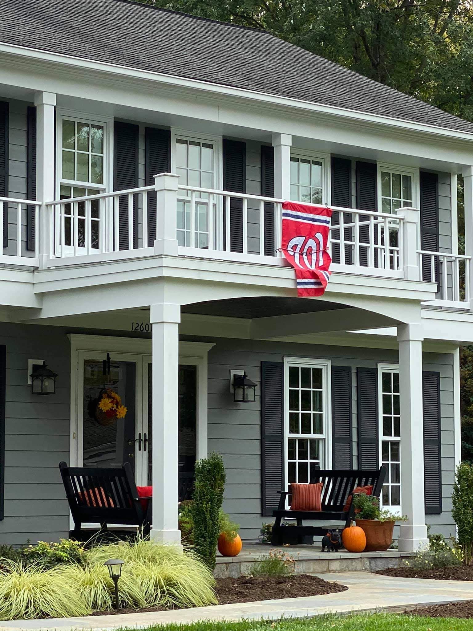 Two-story gray house with white trim, balcony, and red banner, with pumpkins on the front porch.