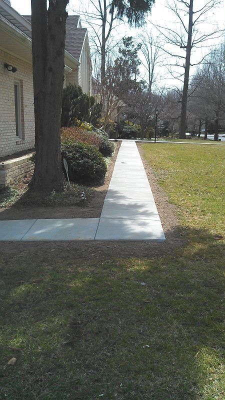 Concrete sidewalk beside a building and tree, with grass and leafless trees in the background.