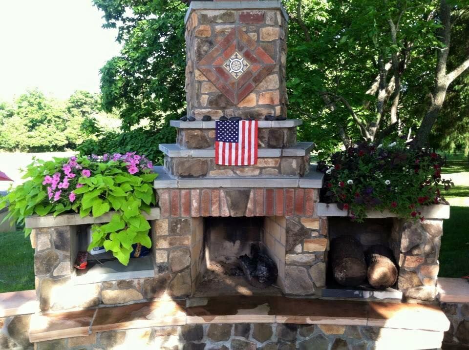 Stone outdoor fireplace with colorful potted flowers and a small American flag in a garden setting