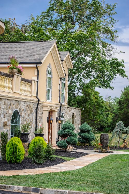 Large stone-and-beige suburban house with landscaped front yard, walkway, and trimmed shrubs under a blue sky