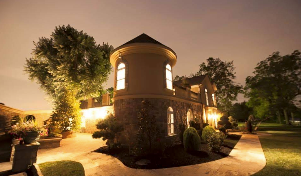 Warmly lit stone house with a turret at dusk, surrounded by trees and a landscaped yard