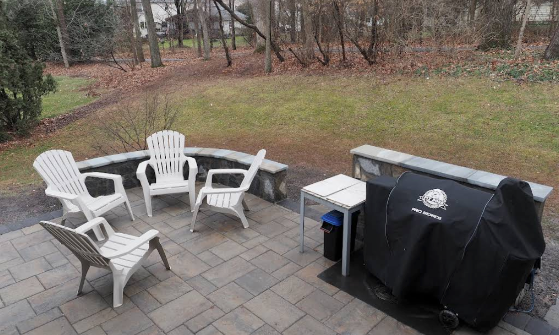 Patio with white chairs around a fire pit beside a covered grill, overlooking a grassy yard and trees