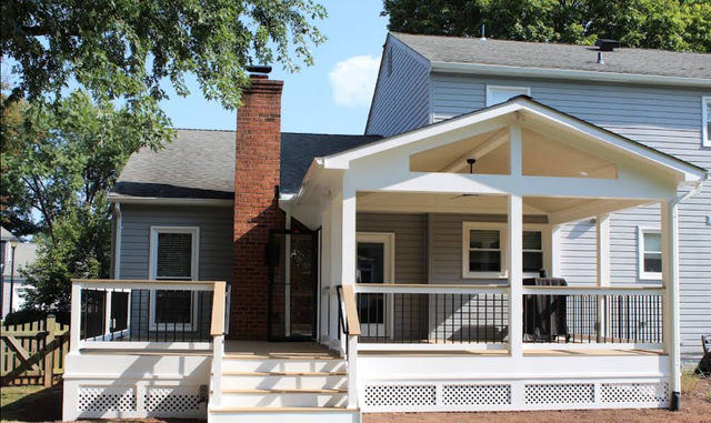 White house with a front porch, steps, brick chimney, and railing under leafy trees.