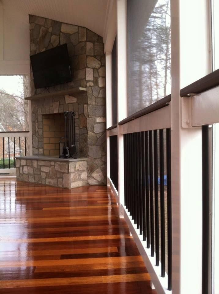 Wooden porch with glossy floor, stone fireplace, and black railing overlooking trees