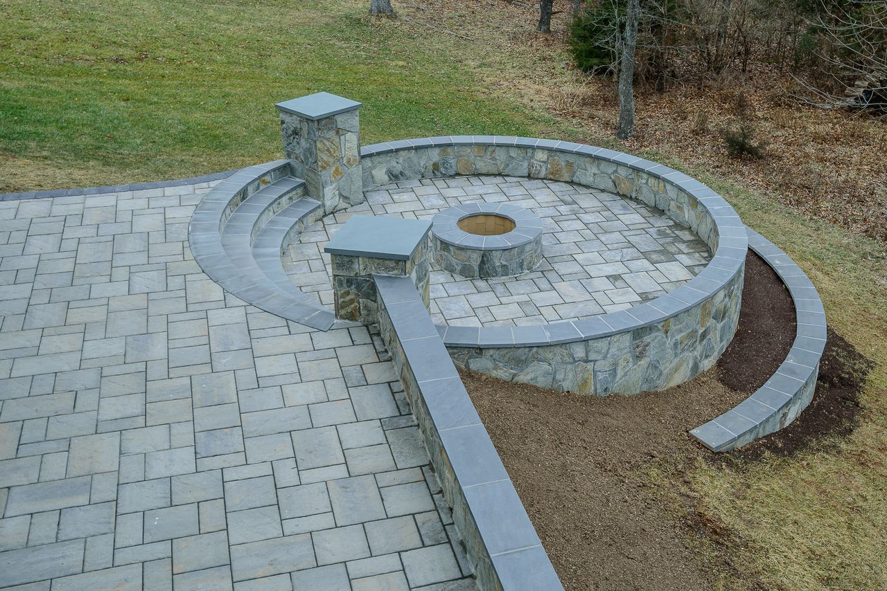 Curved stone bench and circular paved seating area in a park, with a central fire pit and nearby walkway.