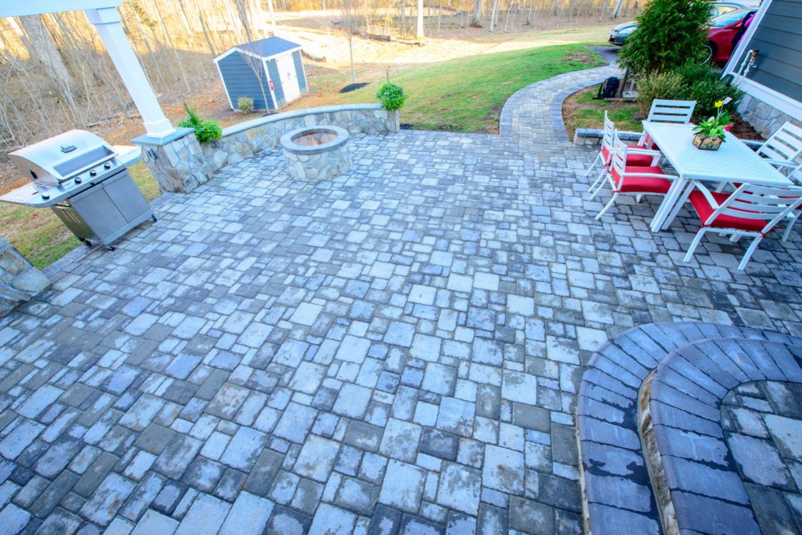 Stone patio with a curved walkway, outdoor grill, and red-and-white dining table set beside a grassy yard