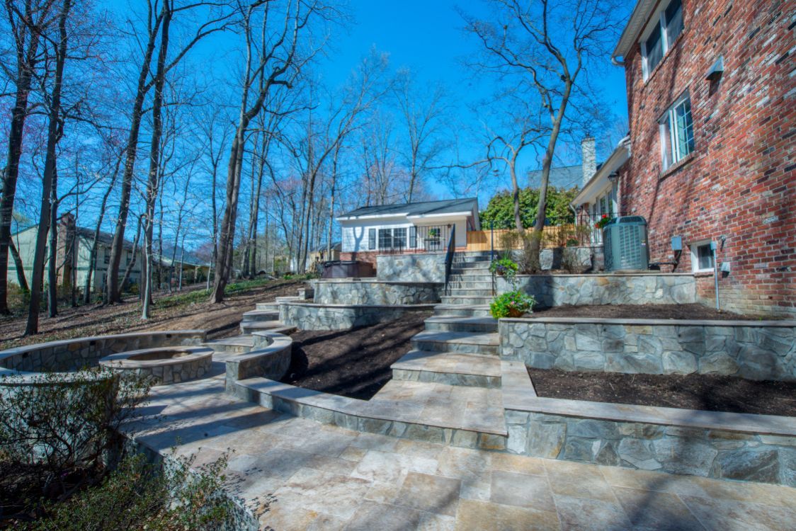 Stone terraced steps and retaining walls leading to a brick house in a wooded yard under a bright blue sky