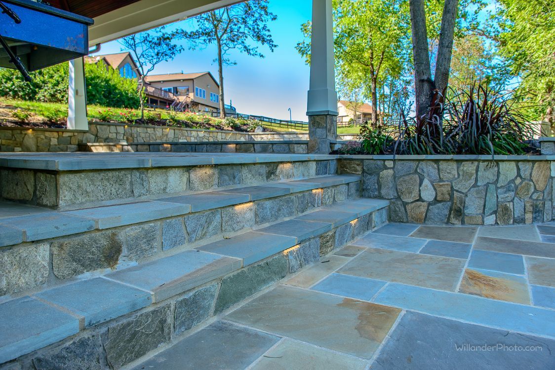 Stone patio steps and a stone retaining wall in a sunny outdoor yard with a house in the background