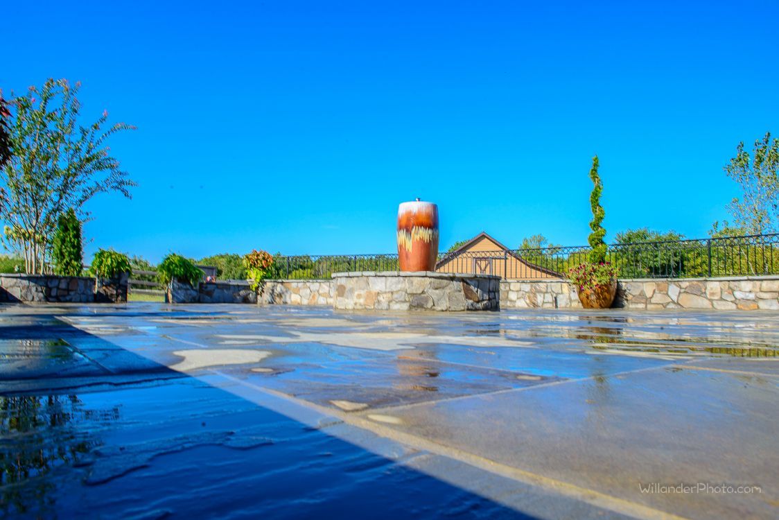 Sunlit wet courtyard with a brick silo, stone wall, and trees under a bright blue sky
