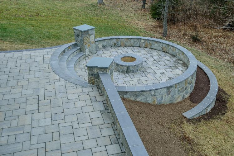 Curved stone amphitheater-style seating with a central circular fire pit beside a paved walkway