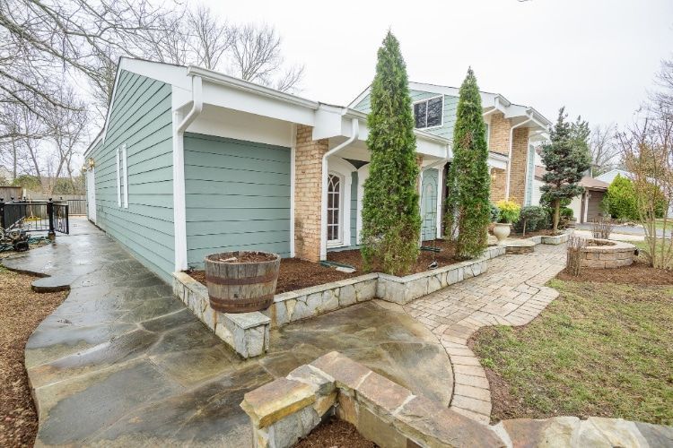 Small house exterior with light blue siding, stone walkway, and leafless landscaping on a wet day