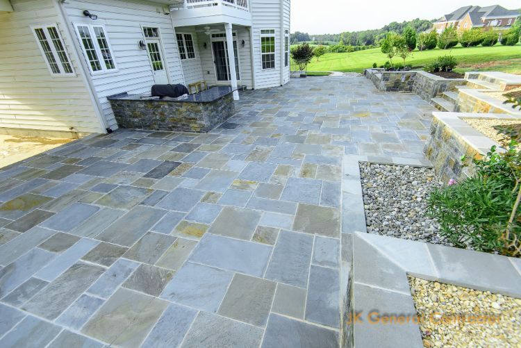 Stone patio and walkway beside a white house, bordered by landscaping and a grassy yard.