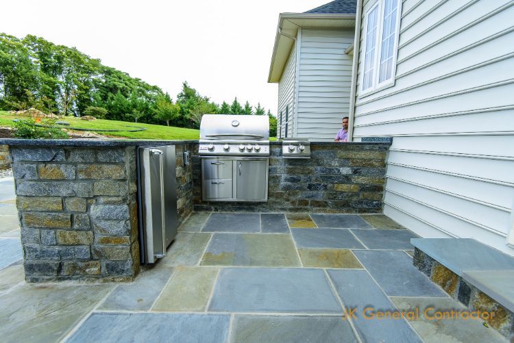 Backyard stone patio with built-in grill beside a white house and landscaped lawn
