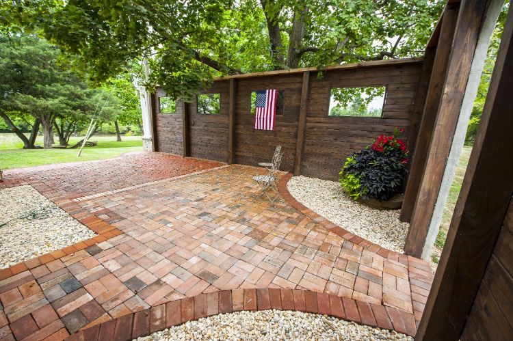 Brick patio with a curved walkway, brick wall, American flag, and flower pot under trees