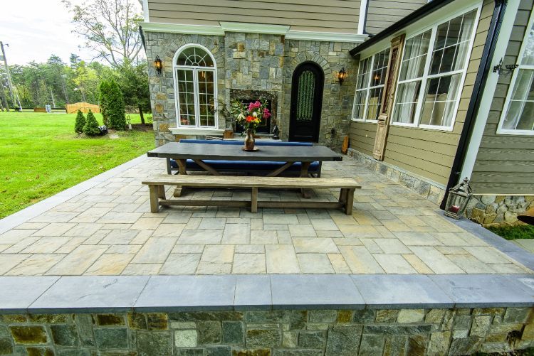 Stone patio with a picnic table and bench beside a house, bordered by a low stone wall and green lawn
