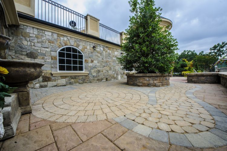 Stone patio with circular paving, stone wall, arched window, and large planters under cloudy sky