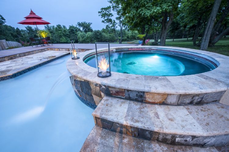 Stone hot tub beside a turquoise pool at dusk, with lanterns and a red-roofed pavilion in the background