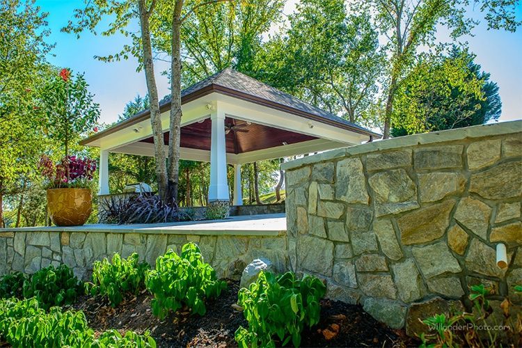 Landscaped garden with stone wall, pavilion, and lush green trees under a bright sky