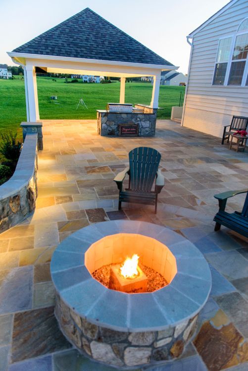 Backyard patio with stone fire pit, chairs, and a lit gazebo overlooking a grassy yard
