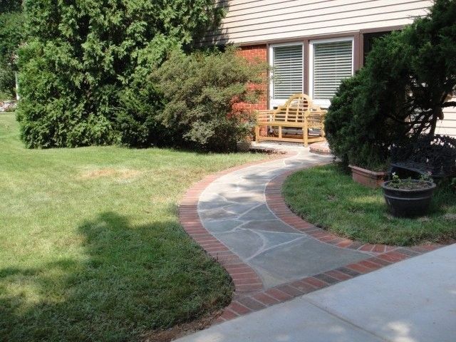 Curved brick-and-stone walkway leading to a wooden bench beside a house and lawn