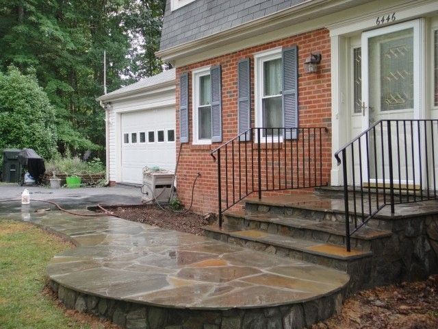 Front entrance of a brick house with stone steps, black railings, and a wet driveway beside a garage