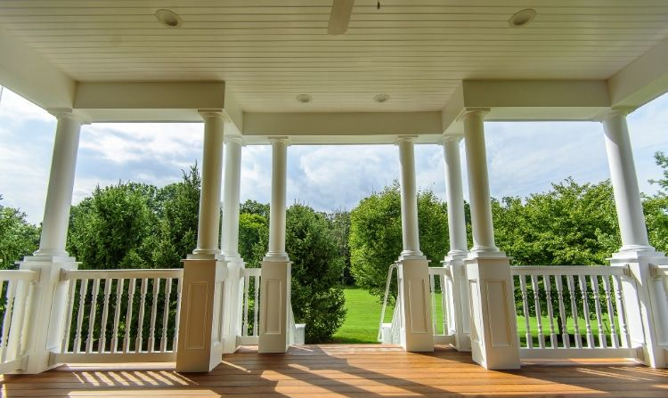 White porch with columns and railings overlooking a green yard under a bright sky