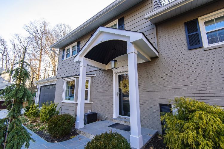 Front entrance of a beige house with white porch columns and trim, surrounded by shrubs and trees.