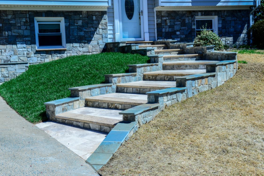 Stone stairway with concrete steps leading up to a house, bordered by grass and dry soil.