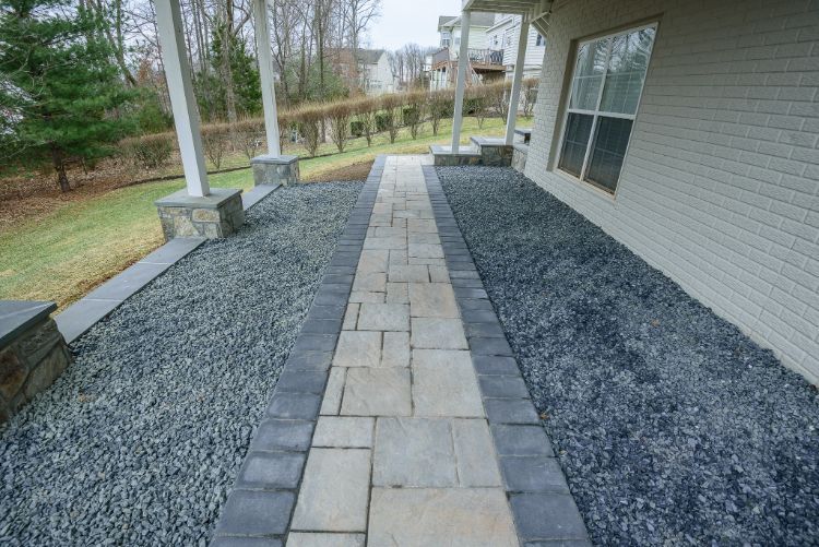 Stone walkway beside a house, bordered by dark gravel landscaping and a covered porch.