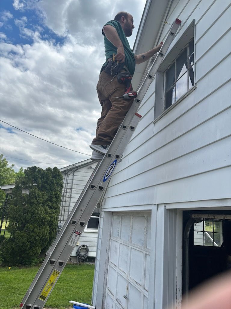 Person on a ladder painting white house trim beside garage door under cloudy sky