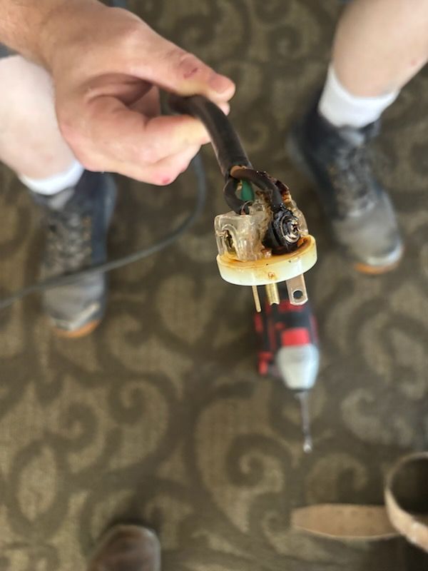 Hand holding a burnt electrical plug with exposed wires and a melted connector on a carpeted floor