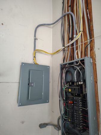 Open electrical breaker panel with exposed wiring on a wall and a second closed gray panel beside it