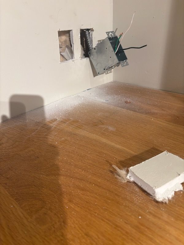 Damaged wall outlet with exposed wires above a wooden floor, broken outlet cover nearby