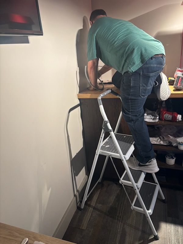 Person standing on a step ladder, reaching over a shelf in a small room with a TV on the wall.
