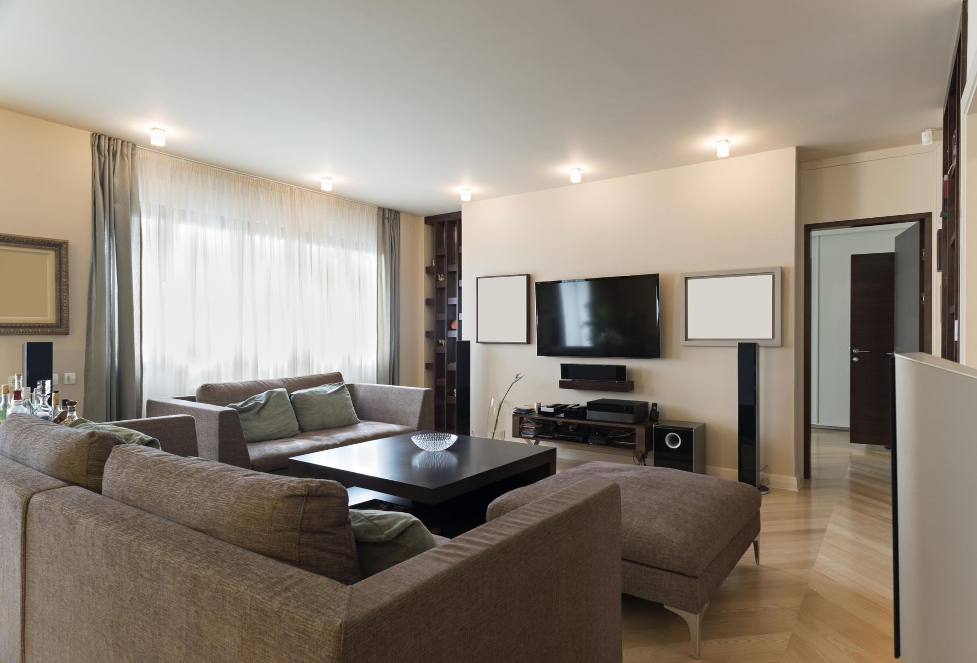 Bright living room with gray sofas, large TV, white walls, and sheer curtains by a window.
