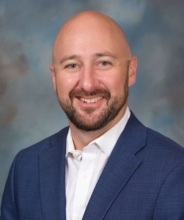A bald man with a beard is wearing a blue suit and white shirt.
