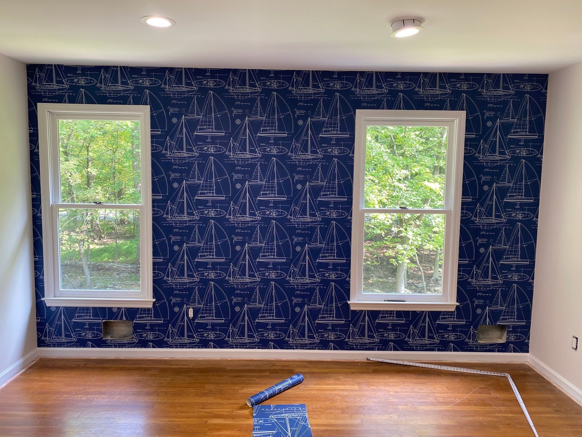 Room with blue nautical wallpaper, two windows, and wooden floor.