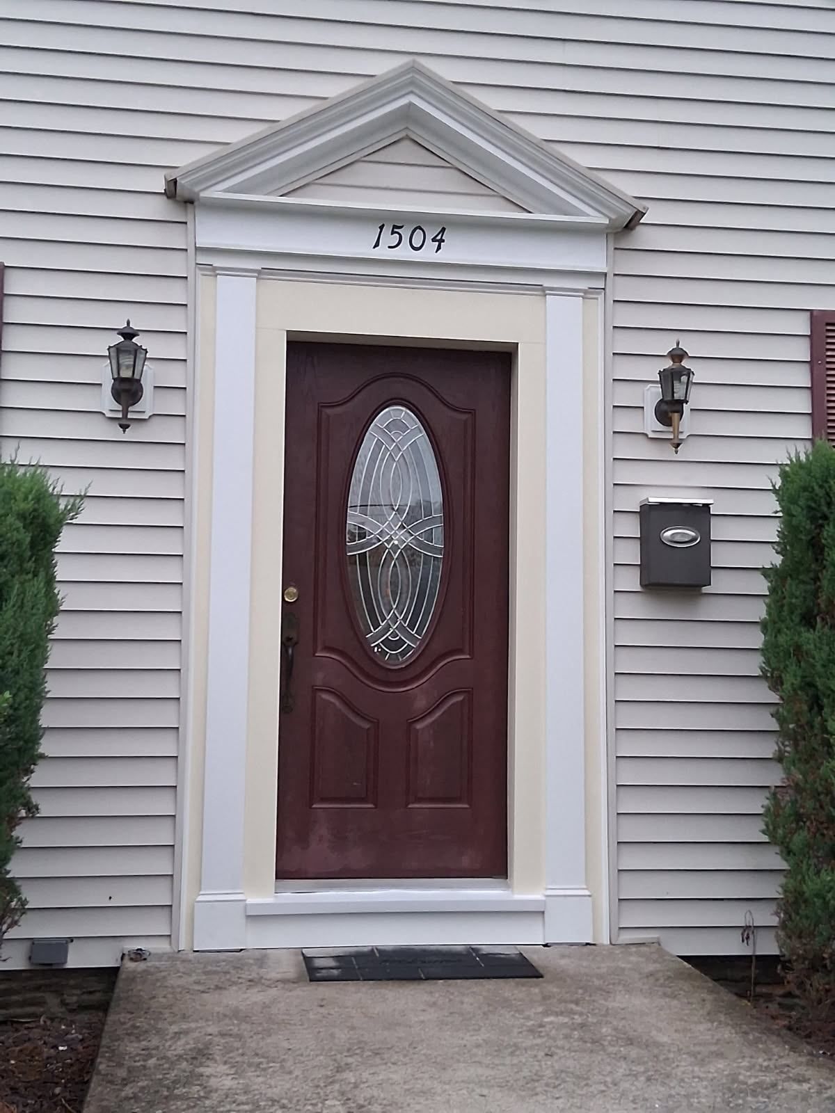 A brown front door with an oval glass insert, framed by white molding and a pediment, labeled 1504 on a house exterior.