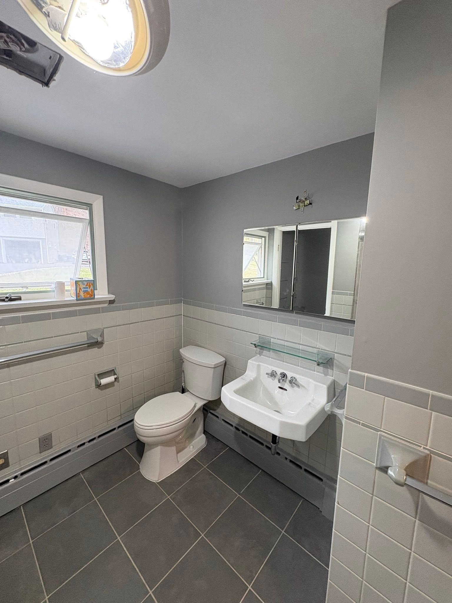 A bathroom featuring gray walls, a white toilet, a wall-mounted sink with a mirror, and tiled walls and floors.