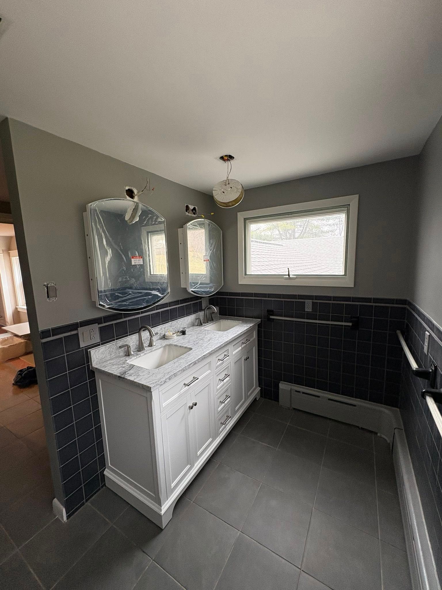 A bathroom with a white double vanity, gray walls, black tiled wainscoting, a window, and two framed mirrors.