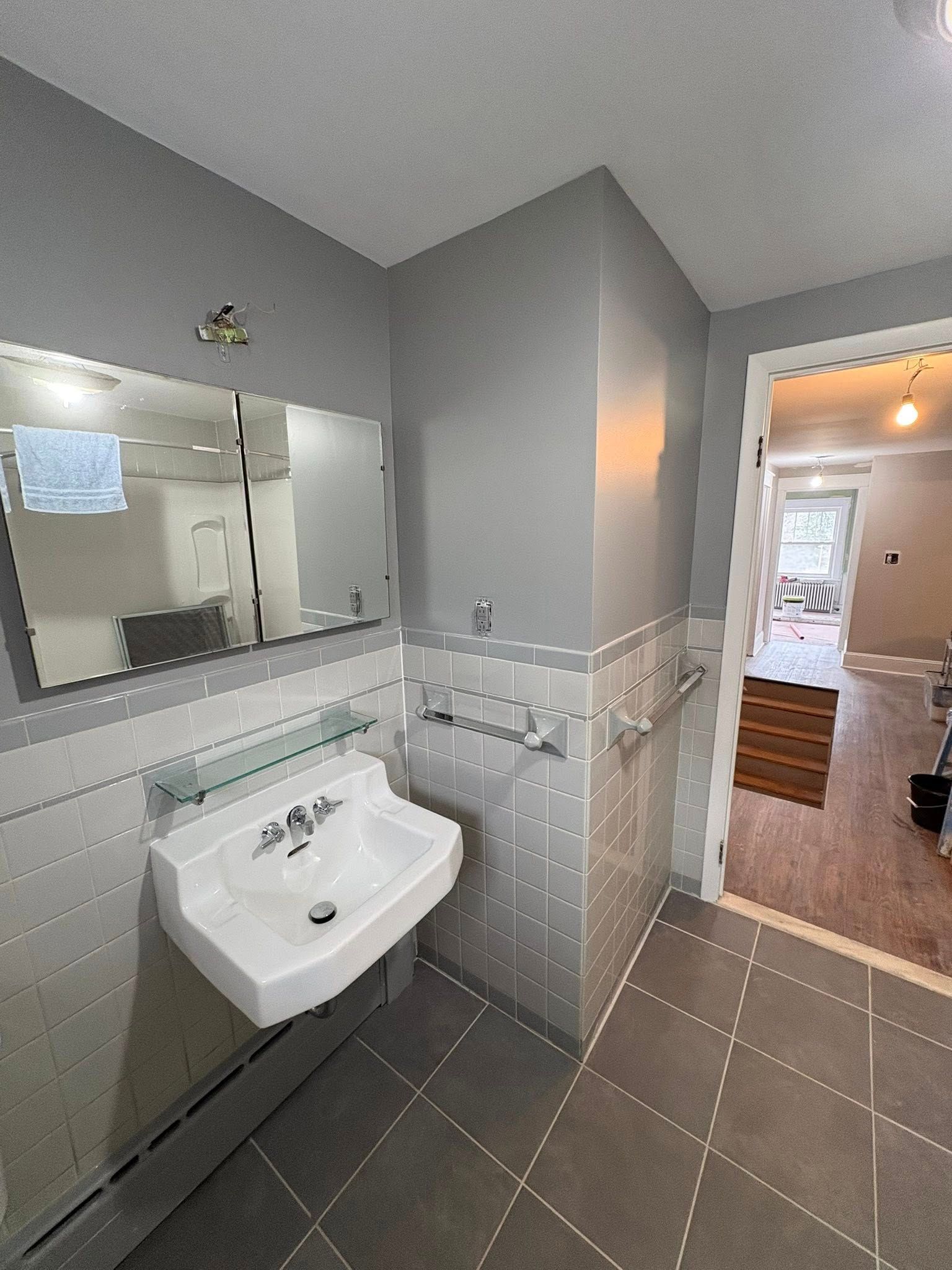 A modern bathroom featuring a white wall-mounted sink, a rectangular mirror, gray tiled walls, and a tiled floor.