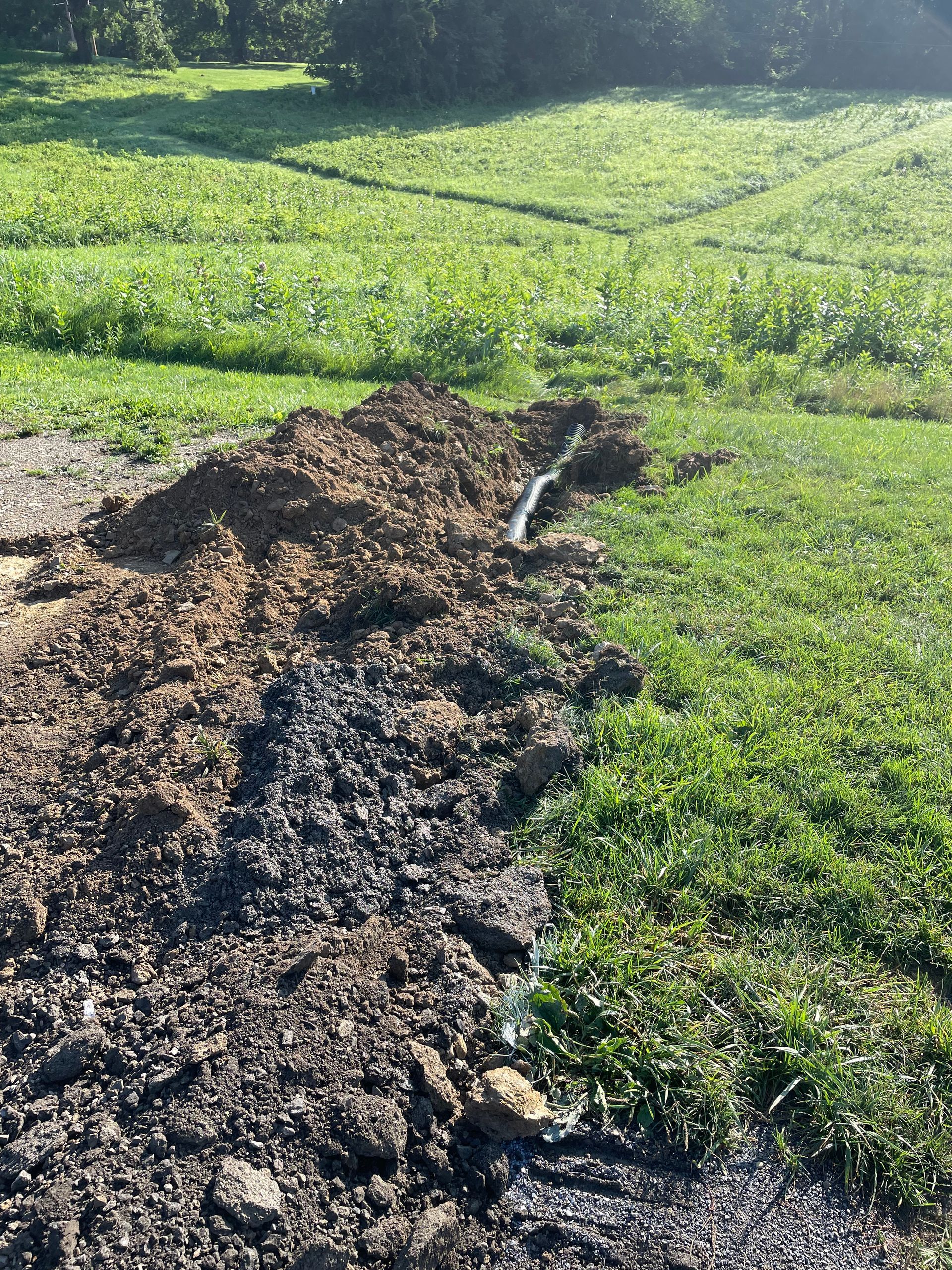 A trench has been dug in a grassy field, exposing a dark pipe running through a pile of displaced soil and mud.