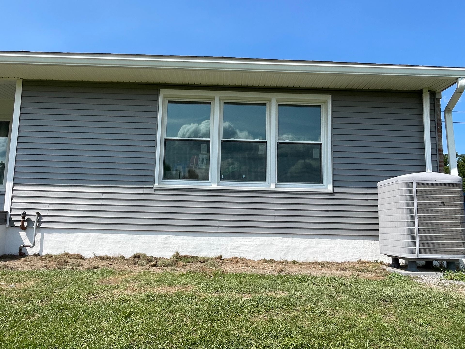 A gray, single-story house exterior with a large three-pane window, white trim, and an outdoor HVAC unit on the right.