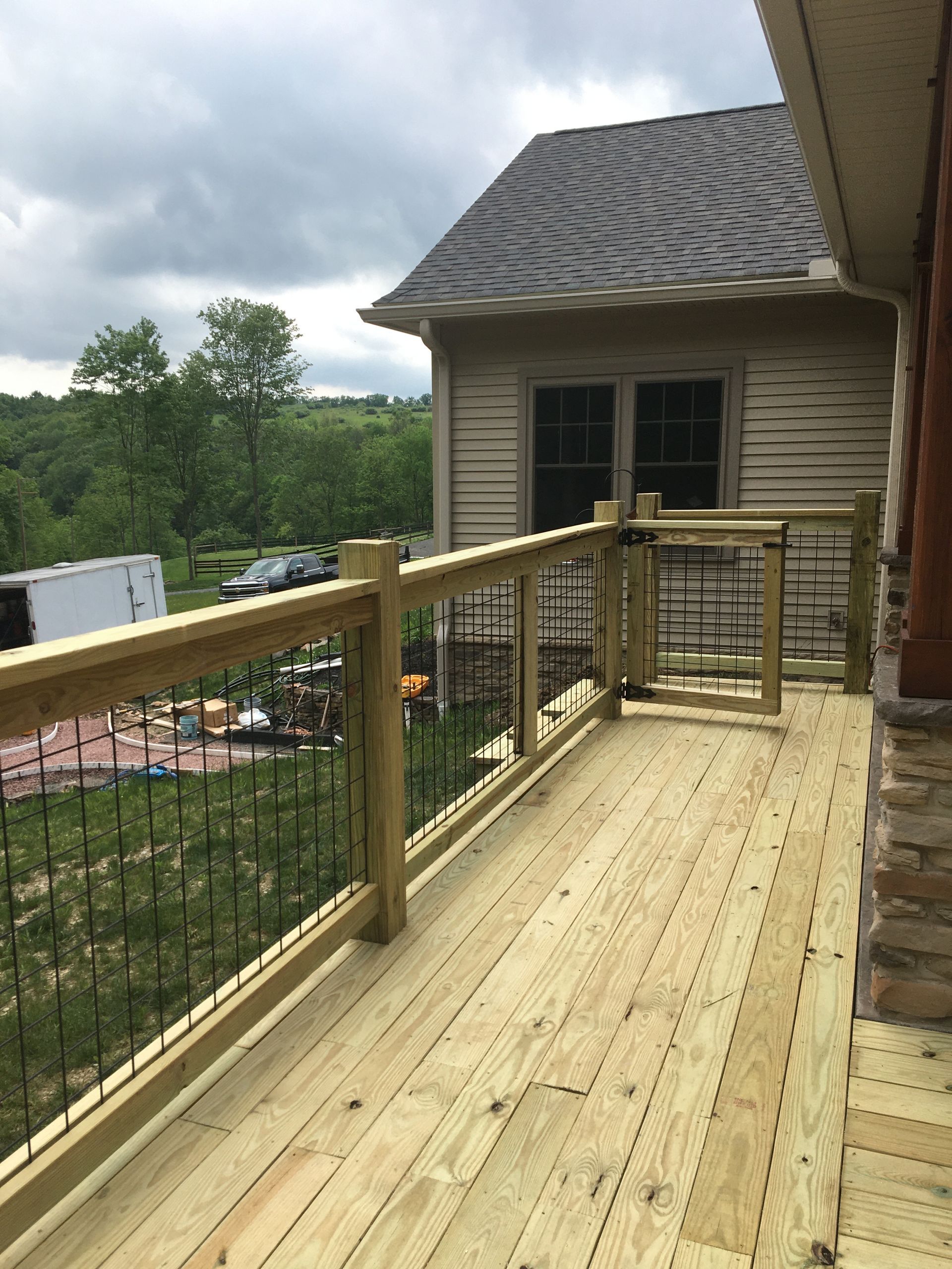 A newly constructed wooden deck featuring a railing with metal balusters and a gate, overlooking a wooded backyard.